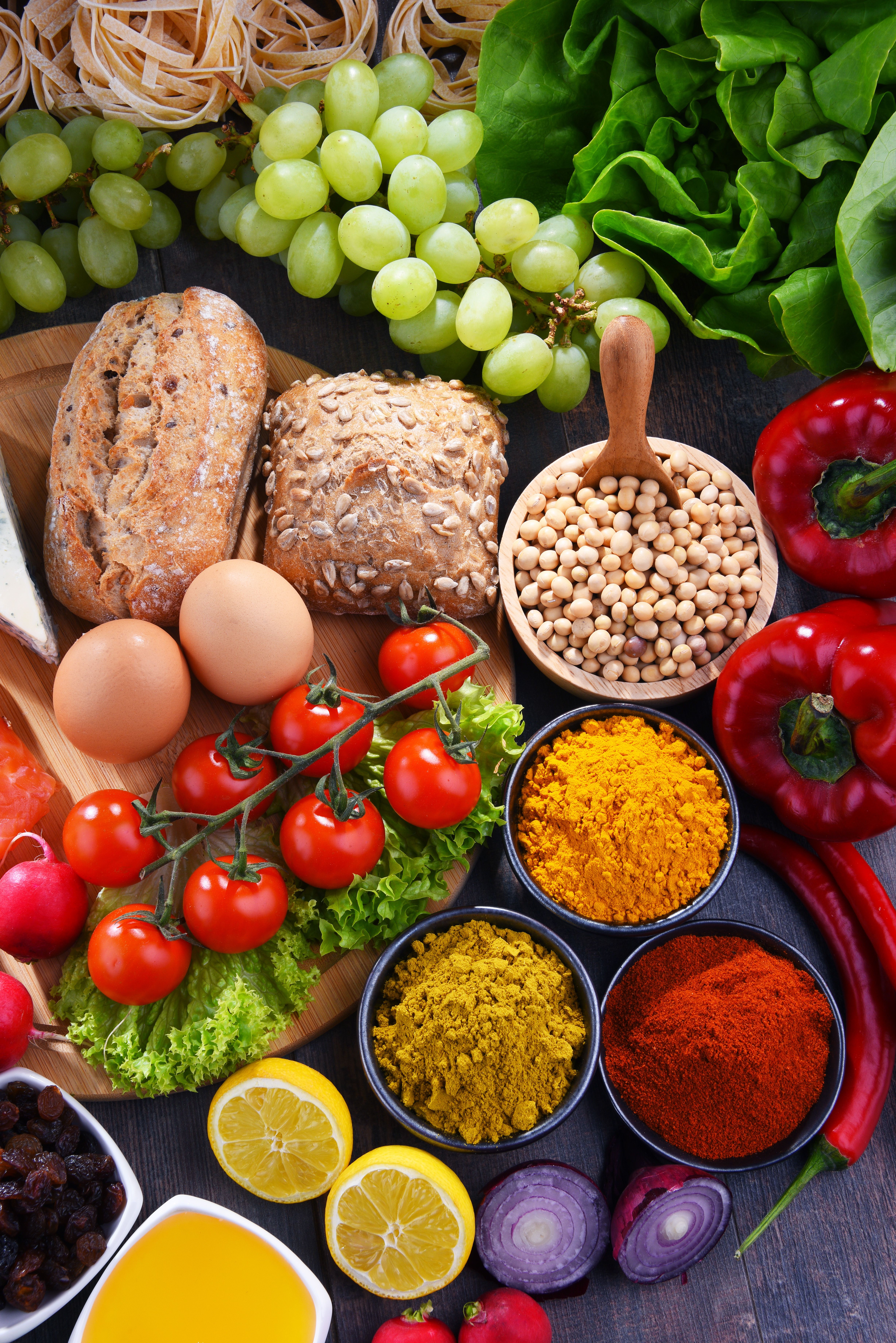 Assorted fresh and healthy foods including whole grain bread, eggs, cherry tomatoes, lettuce, grapes, red peppers, soybeans, spices, lemons, raisins, onion, and olive oil, representing balanced nutrition.
