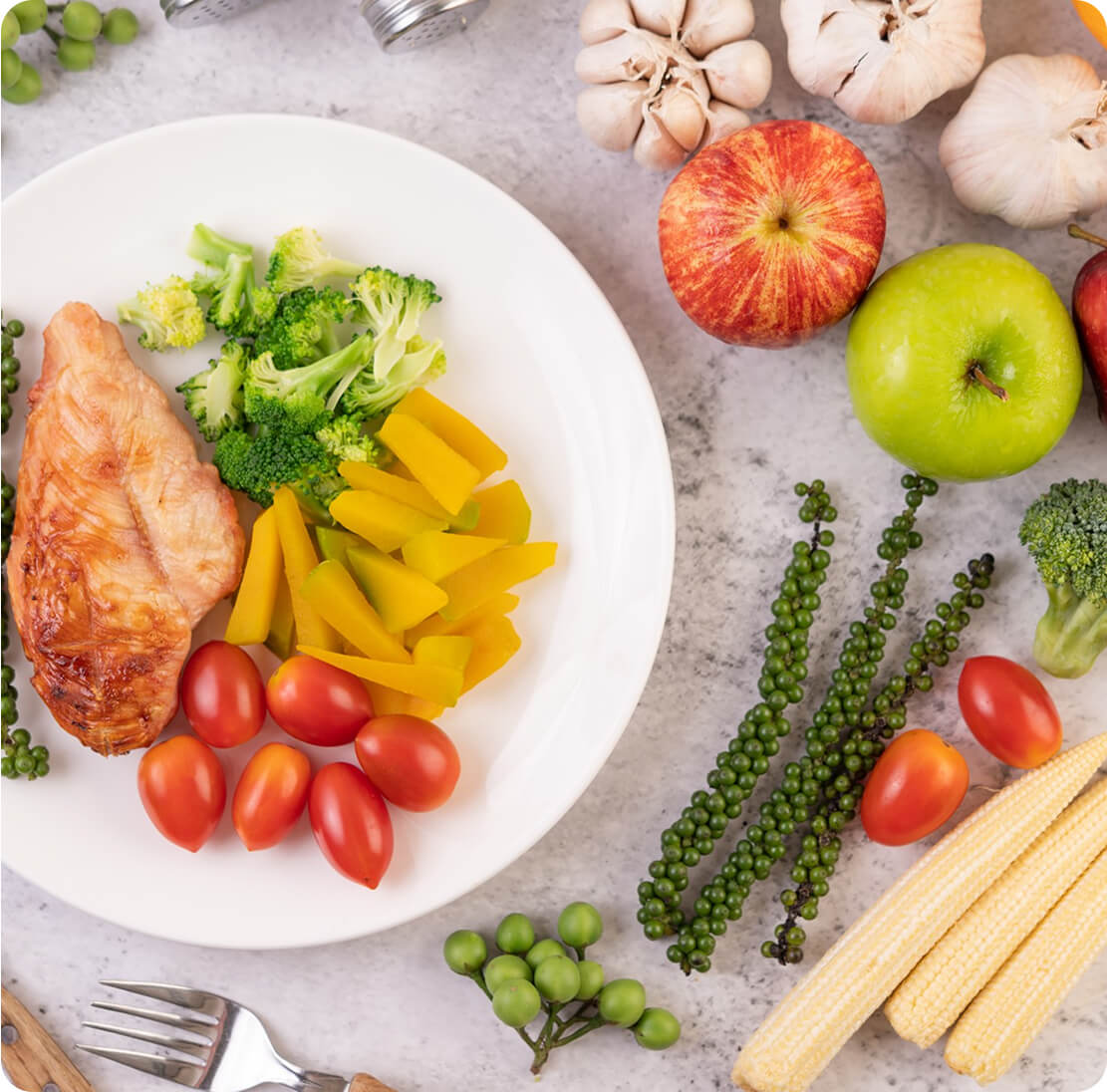 Healthy plate with grilled chicken, broccoli, yellow peppers, and cherry tomatoes, surrounded by apples, garlic, and fresh green peppercorns, representing balanced nutrition.